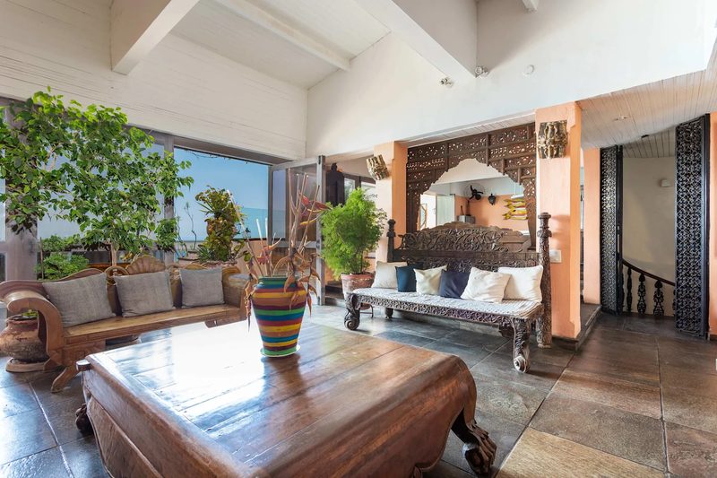 A living room with dark carved wooden benches with cushions, a large low wooden table with a striped vase, stone floor, white beamed ceiling, potted trees and a window open to the sea.
