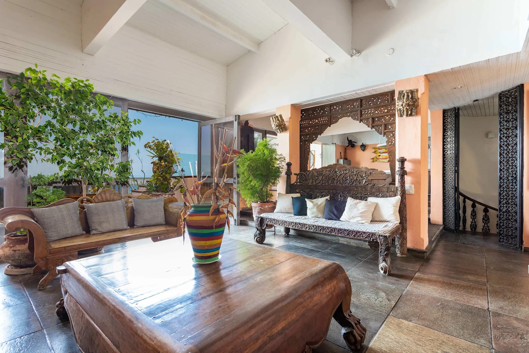 A living room with dark carved wooden benches with cushions, a large low wooden table with a striped vase, stone floor, white beamed ceiling, potted trees and a window open to the sea.