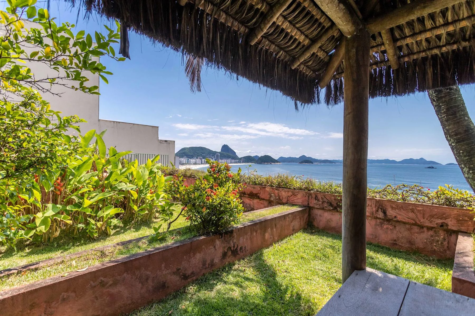 A rooftop garden with a lawn, flowering tropical plants and low pink walls seen from under a thatched shelter, with a bay, a city beachfront and a domed rock peak in the distance.