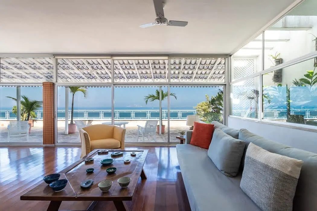 A living room with a grey sofa with cushions, a yellow armchair, a low wooden table set with ceramic bowls and a polished wood floor, glass doors opening to a terrace and the sea.