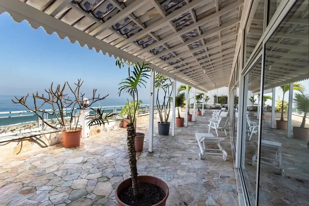 A long covered terrace paved in irregular stone, with potted palms, white wooden loungers and chairs, a painted tile ceiling and sliding glass doors on the right, open to the sea on the left.