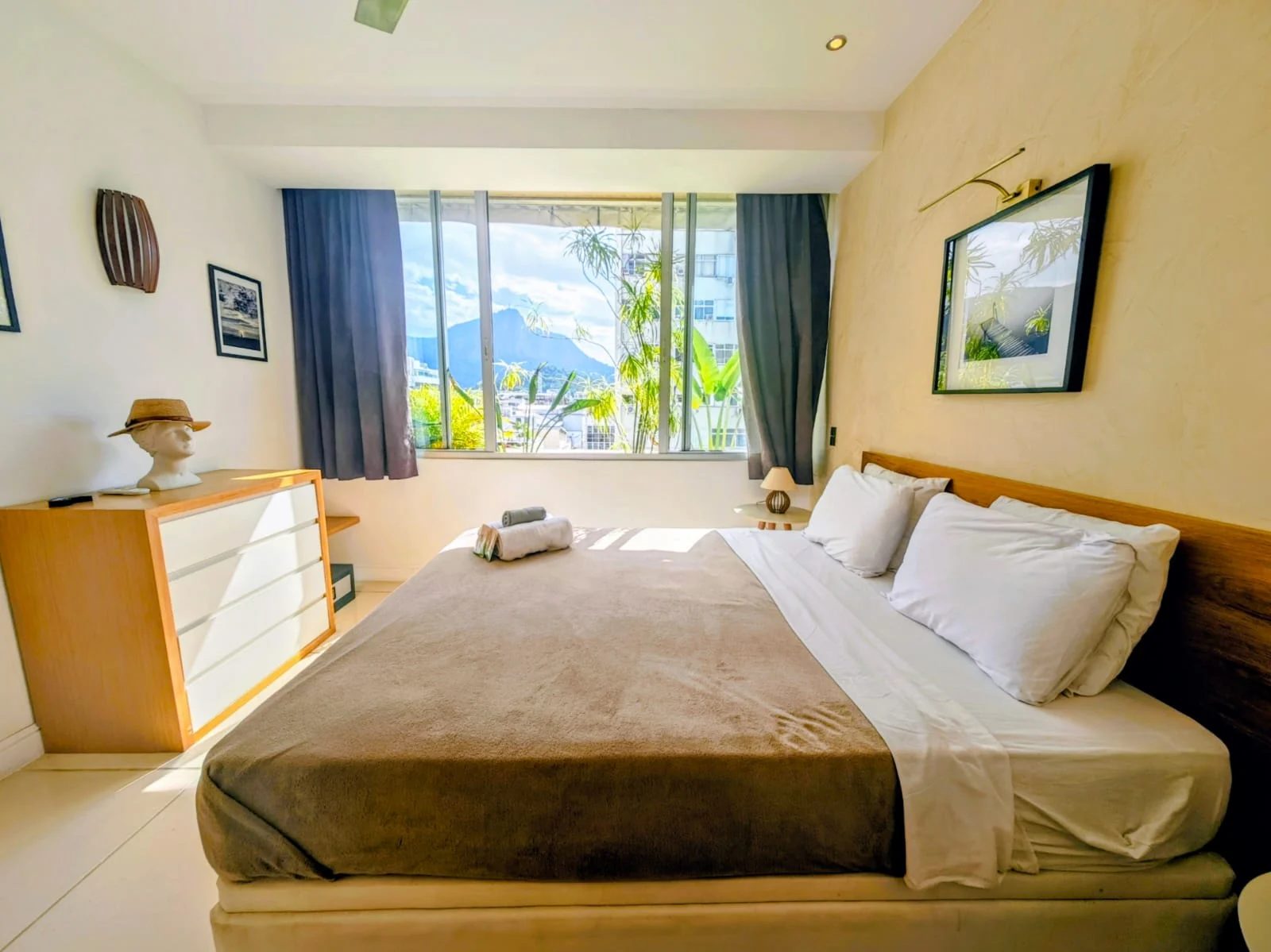 A bedroom with a double bed with a beige cover and white pillows, a wooden chest of drawers with a bust wearing a straw hat, and a window with a view of a mountain.