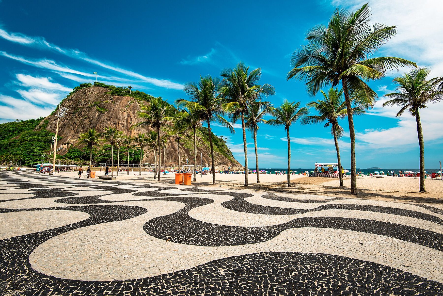 A promenade paved in black and white stone in a wave pattern, with tall coconut palms, sand, beach umbrellas and a steep rocky hill with a flag on top under a blue sky.