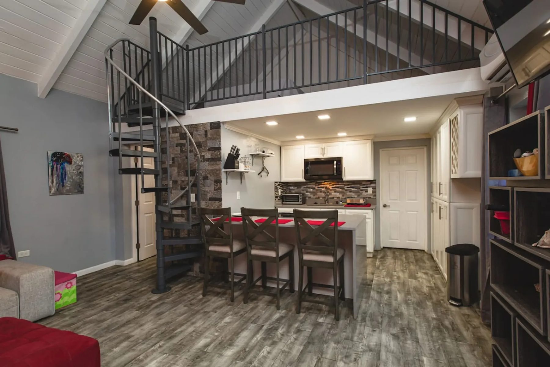 A cottage interior with a grey spiral staircase rising to a railed loft, a small kitchen with white cabinets and a counter with three high chairs beneath it, a grey wood-look floor and a white pitched ceiling.