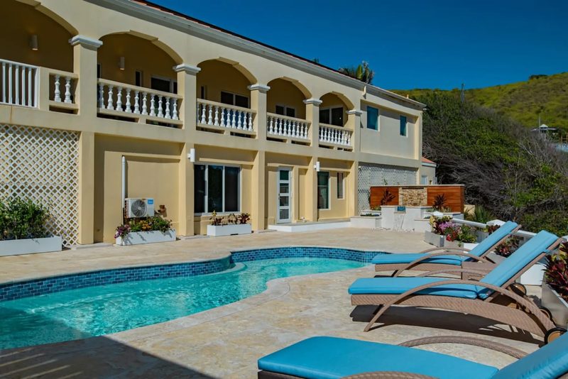 A cream two-storey house front with five arched upper verandas and white balustrades, seen across a curved pool and blue-cushioned sun loungers on a stone deck in bright daylight.
