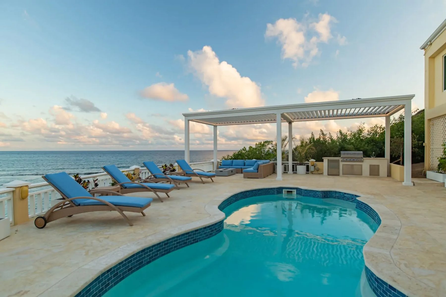 A curved pool on a pale stone deck with four blue sun loungers, a white louvred pergola over outdoor sofas and a built-in grill, and the open sea beyond a white railing.