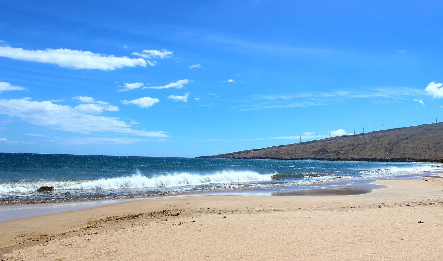 Wide sandy beach with a breaking wave, deep blue sea and sky, and a dry brown mountain slope on the right topped by a line of wind turbines.