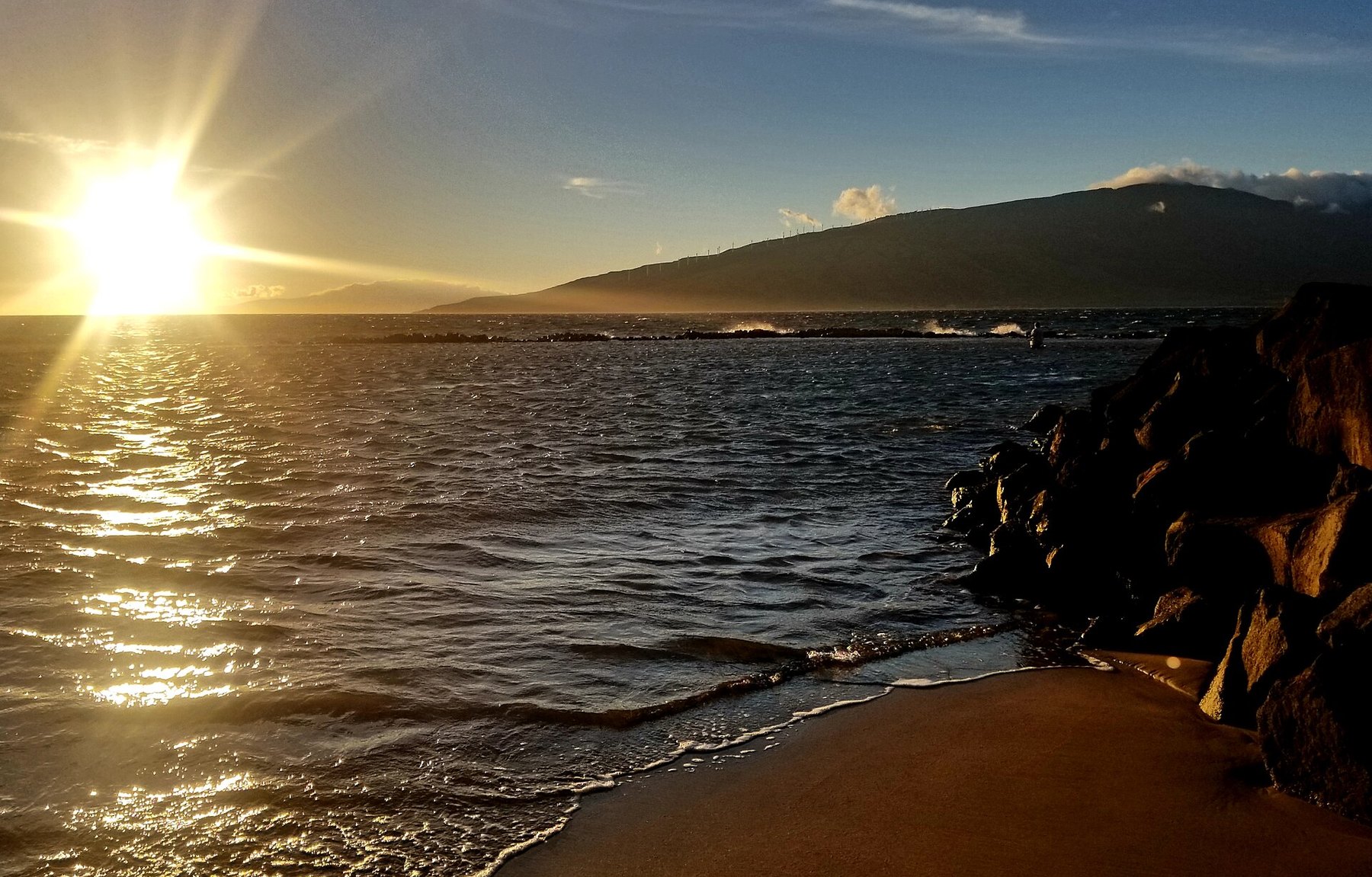 The sun low over calm water at sunset, with a dark lava-rock wall at the right, a strip of sand in the foreground and a dark ridge of hills on the horizon.