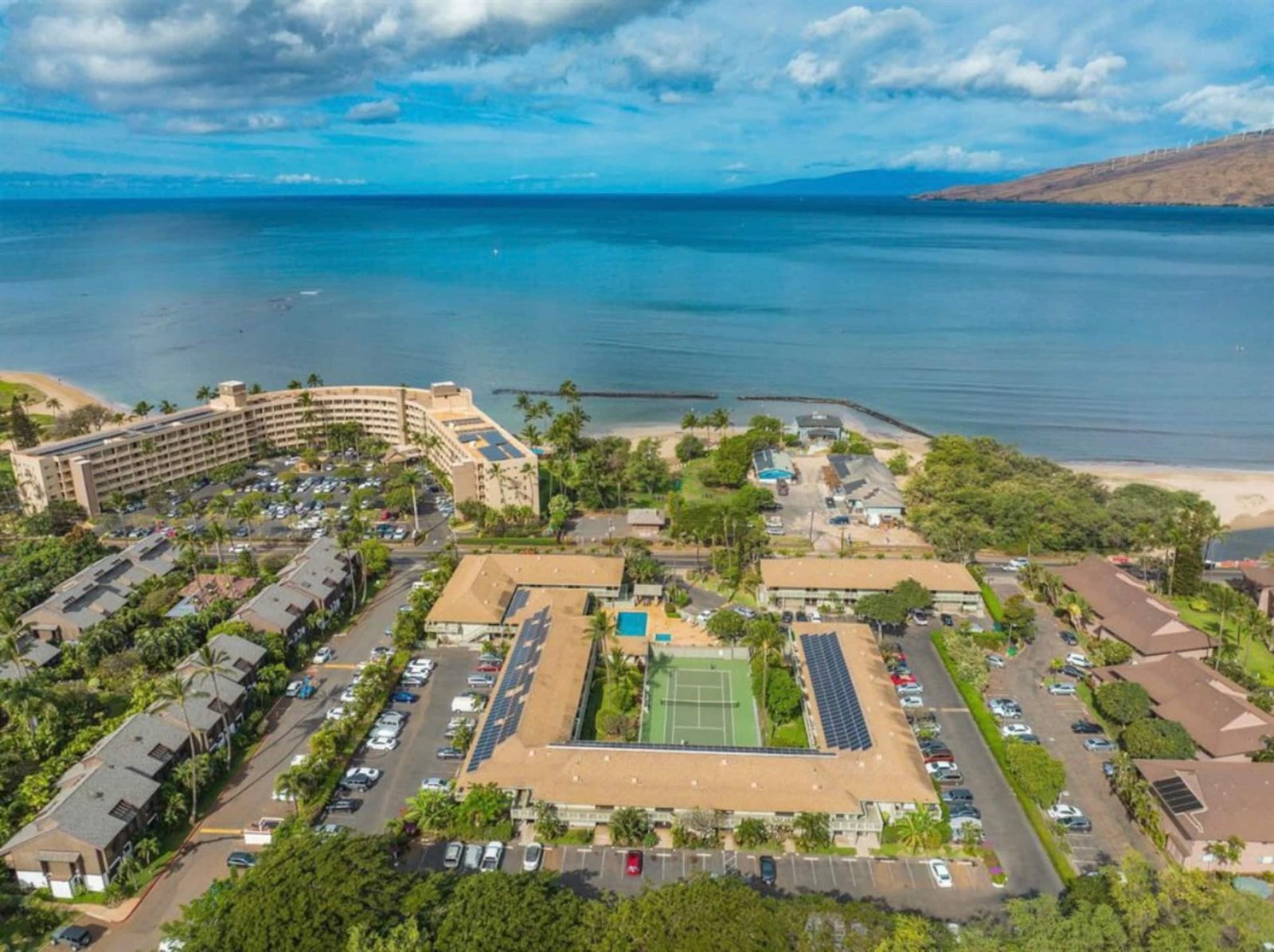 Aerial view of a U-shaped low-rise condominium complex around a tennis court and pool, with car parks, a road, a curved mid-rise building on the shore, rock walls in the shallows and a mountain slope with wind turbines.