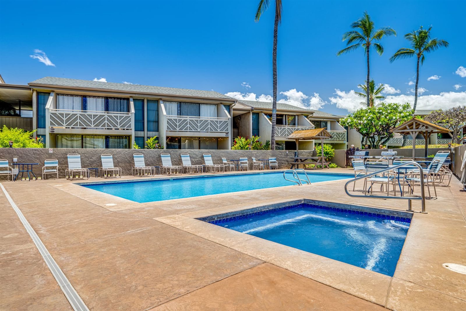Swimming pool and a square hot tub on a tan concrete deck lined with white loungers, backed by two-storey condominium buildings with patterned balcony railings, a barbecue shelter and tall palms.