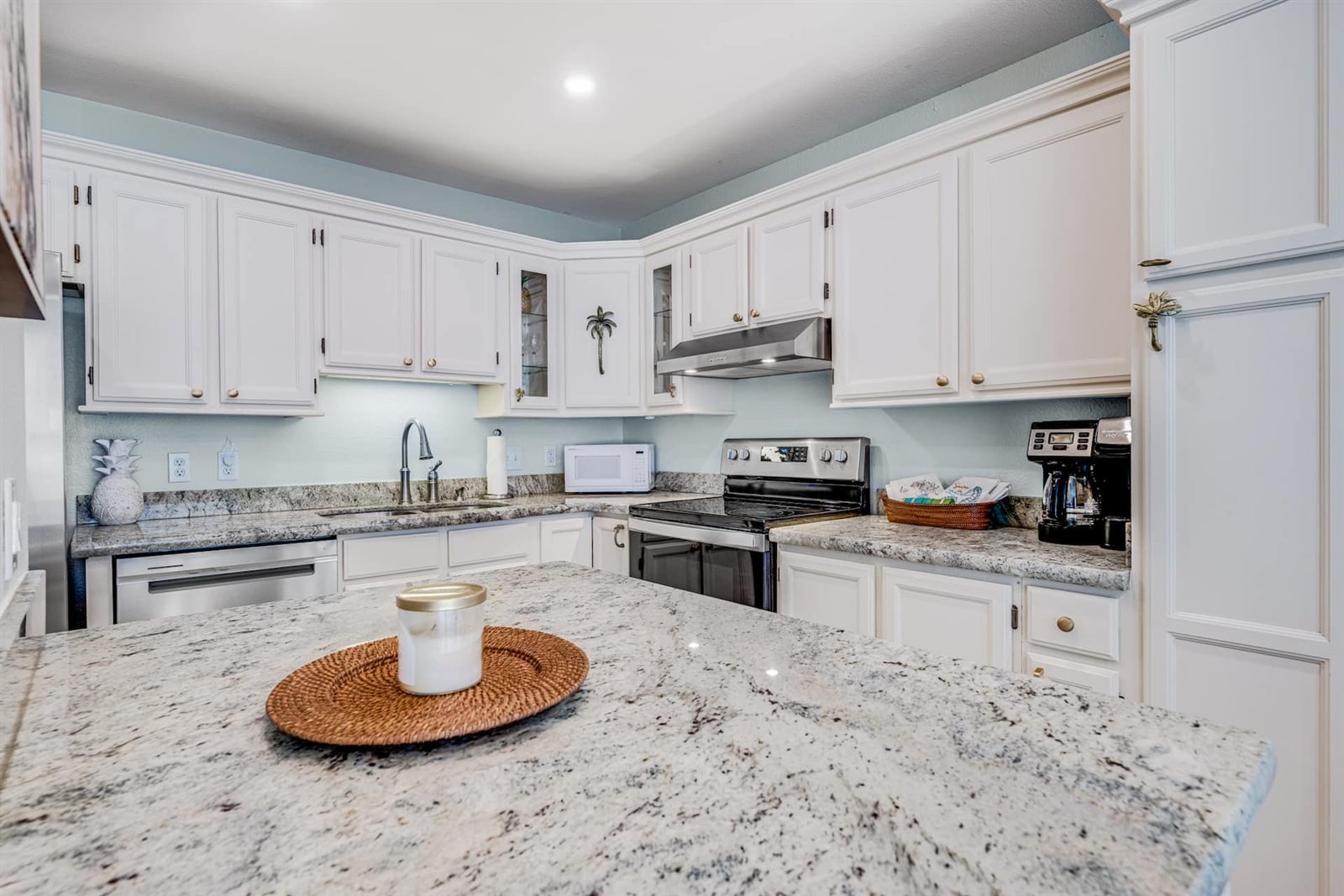 Kitchen with white raised-panel cabinets, pale blue walls, speckled granite counters, a stainless range, hood and dishwasher, a white microwave and a coffee maker, seen across a granite peninsula.