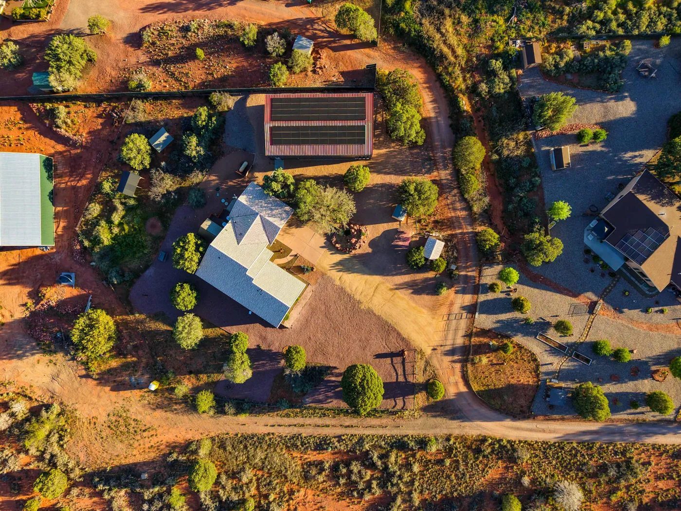 Straight-down drone view of a rural lot: an L-shaped house with a pale roof among juniper trees, a long building with a red-trimmed roof covered in solar panels just behind it, dirt drives, red soil and neighbouring lots.