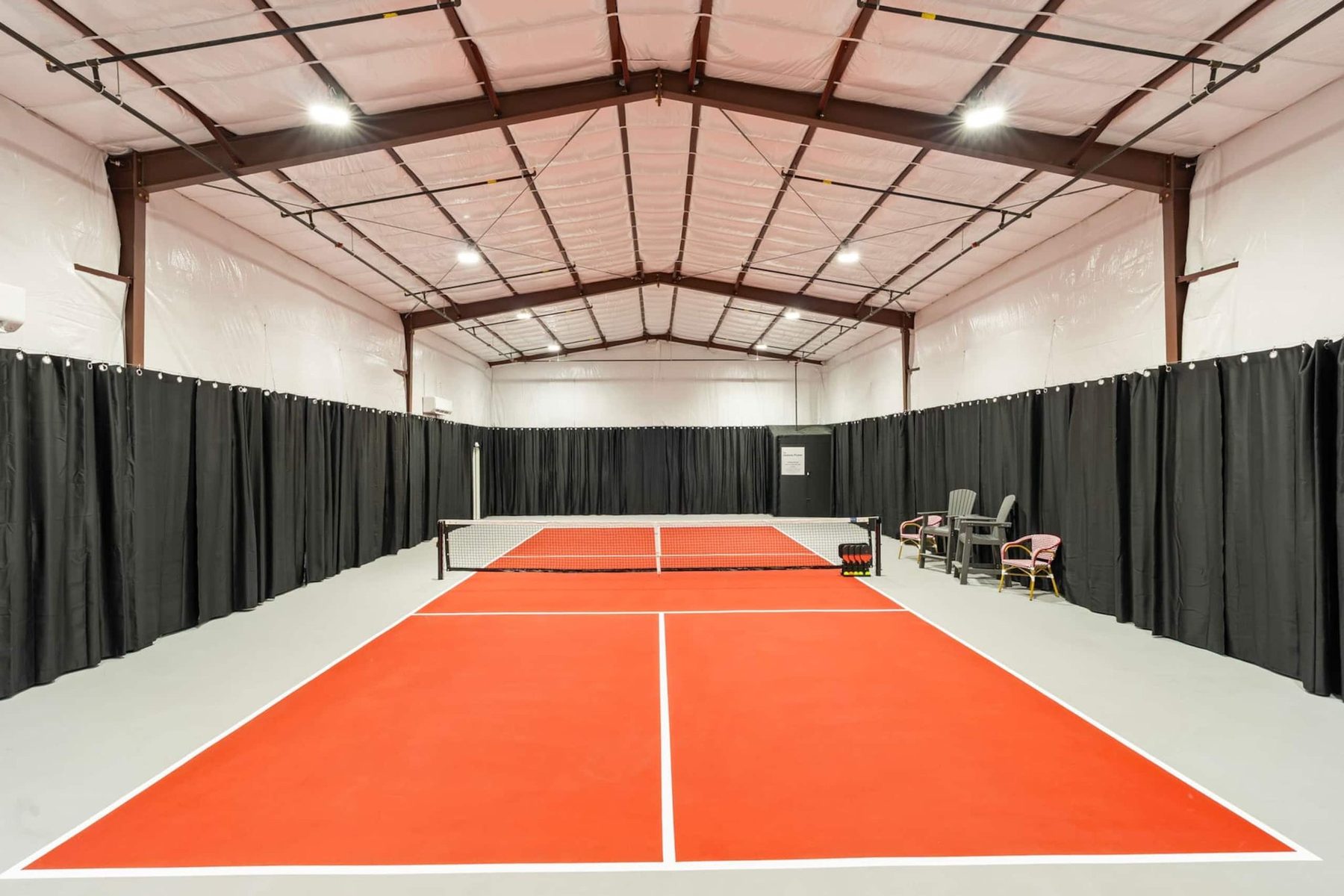 An empty indoor pickleball court seen down its centre line: an orange playing surface with white lines on a grey floor, a net across the middle, black curtains along the walls and a steel-framed roof with lights.