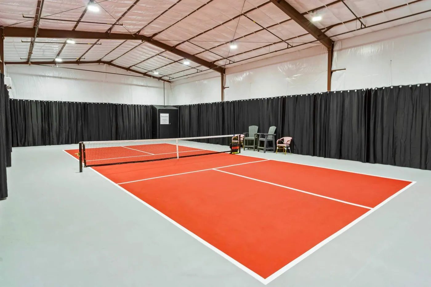 The indoor pickleball court seen from one corner: an orange court and net on a grey floor, black curtains on every side, three chairs beside the net post and an insulated steel-framed roof with lights.