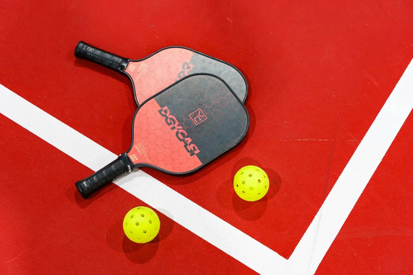 Two red-and-black pickleball paddles and two yellow perforated balls lie on a red court surface where two white lines meet, photographed from above.