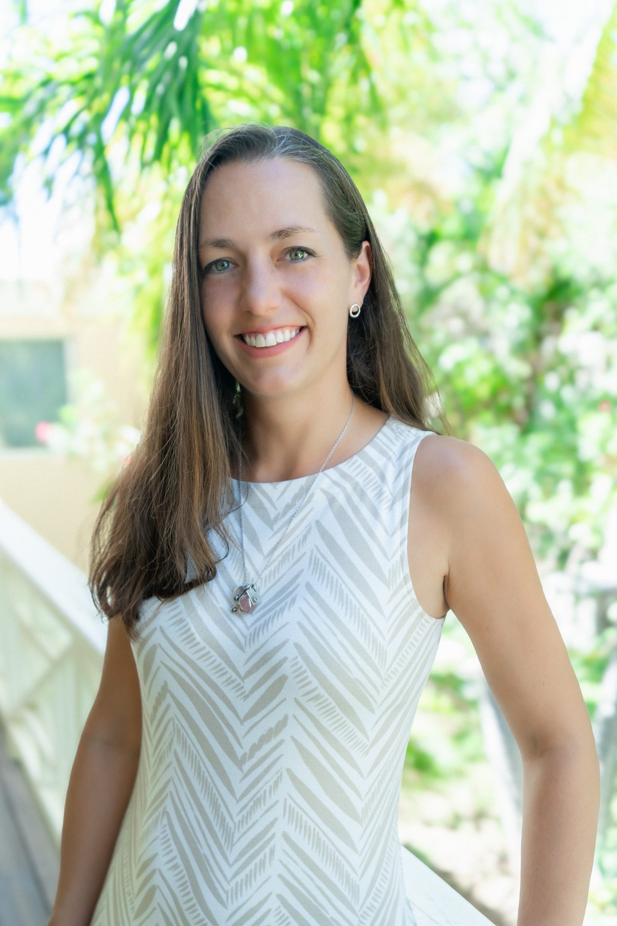 A portrait of a smiling woman with long brown hair wearing a sleeveless white and grey patterned dress, standing on a walkway in front of sunlit tropical greenery.