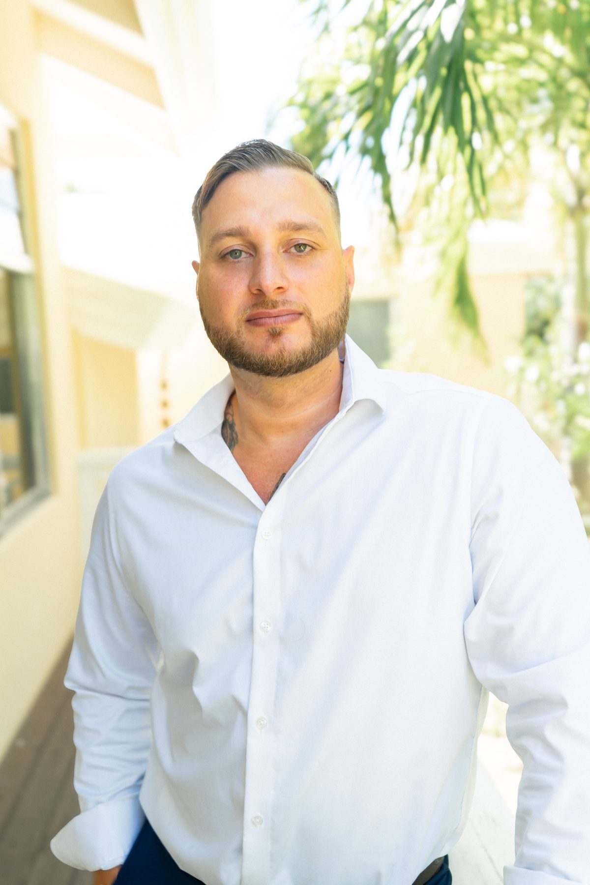 A portrait of a man with short hair and a trimmed beard wearing a white button-down shirt, standing on a bright walkway beside a pale yellow building with palm fronds behind him.