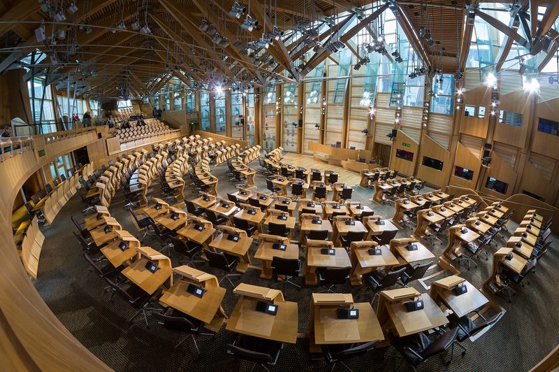 The empty debating chamber of the Scottish Parliament, with curved rows of wooden desks under a timber-beamed roof