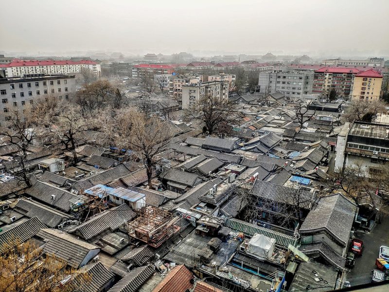Grey tiled roofs of low courtyard houses packed together under a hazy winter sky, with mid-rise apartment blocks behind them