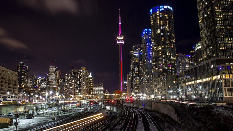 Lit condominium towers and the CN Tower at night above a railway corridor, with light trails from passing trains
