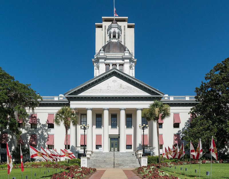 The white, columned old Florida State Capitol with red-striped awnings and a small dome, the tower of the new Capitol rising behind it and state flags along the lawn