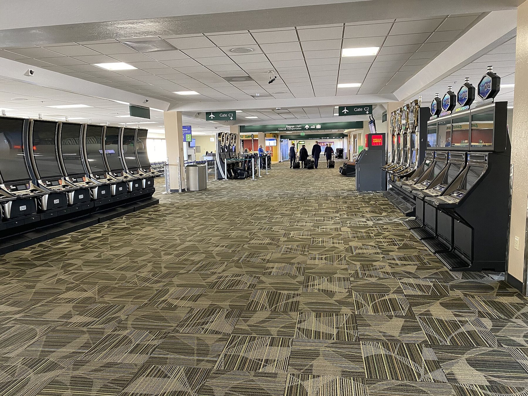 A nearly empty airport concourse lined with rows of slot machines, with a handful of travellers wheeling bags in the distance