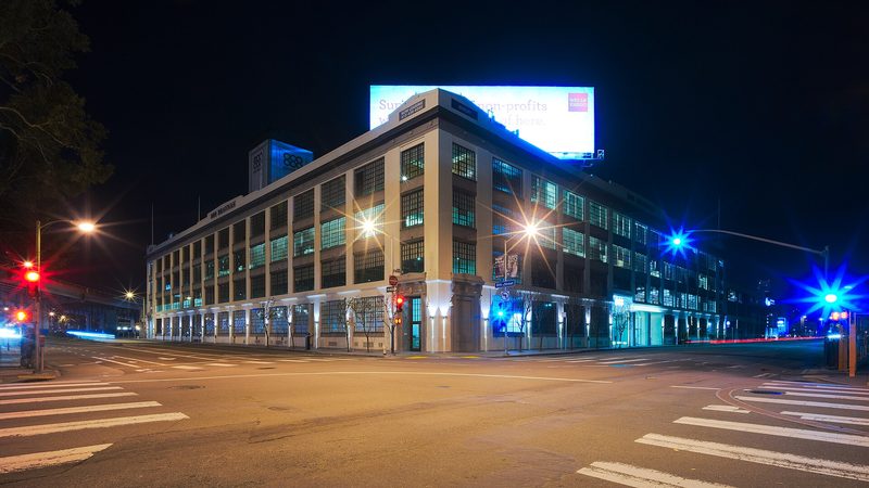 A four-storey cream-coloured building with tall grid windows on an empty street corner at night, with a lit billboard on its roof and traffic lights at the crossing.