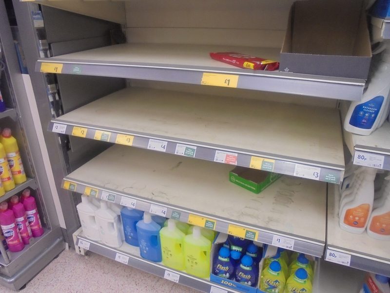 Supermarket shelves in the cleaning products aisle standing largely empty, with price labels along the shelf edges