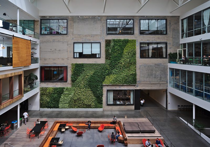 A multi-storey office atrium with a concrete wall partly covered by a green plant wall, open and glass-fronted floors on either side and a sunken seating area with red chairs on the ground floor