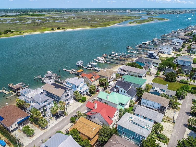 An aerial view of two- and three-storey beach houses with coloured roofs beside a tidal channel, with boat docks along the shore and salt marsh beyond