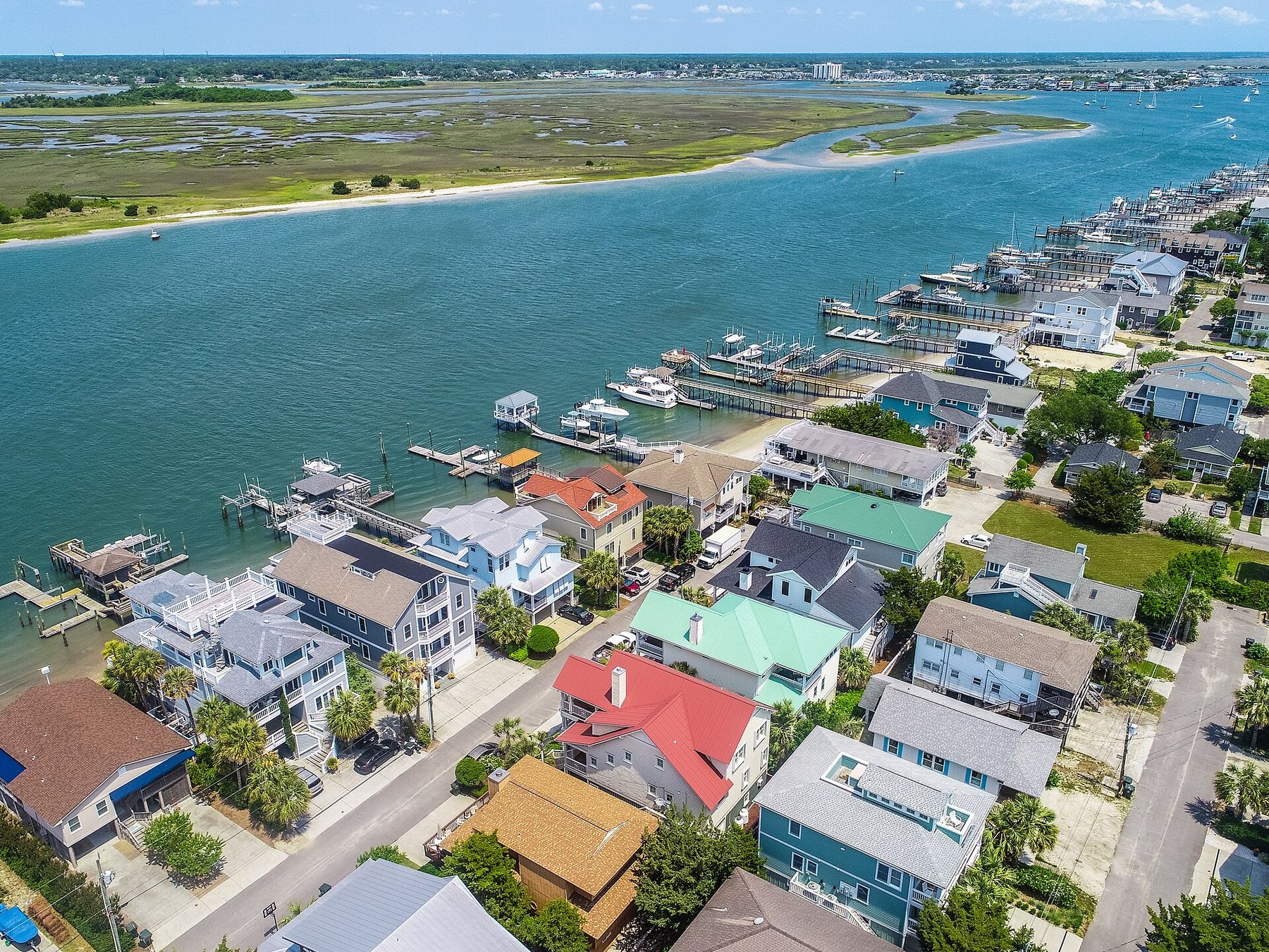An aerial view of two- and three-storey beach houses with coloured roofs beside a tidal channel, with boat docks along the shore and salt marsh beyond