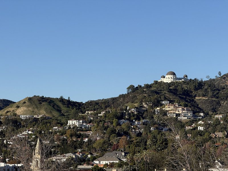 White and terracotta houses are scattered across a green hillside under a clear blue sky, with the domed Griffith Observatory on the ridge above them and a church spire in the foreground.