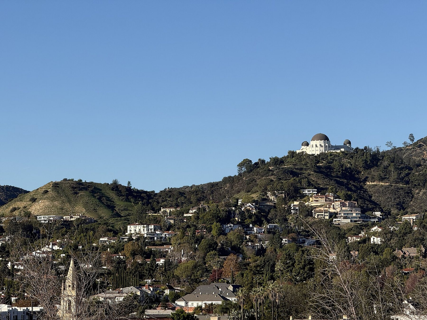 White and terracotta houses are scattered across a green hillside under a clear blue sky, with the domed Griffith Observatory on the ridge above them and a church spire in the foreground.