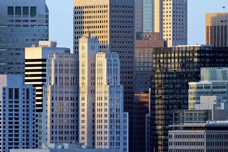 Office towers of glass, concrete and pale stone stand packed closely together in warm evening light, filling the frame from edge to edge.
