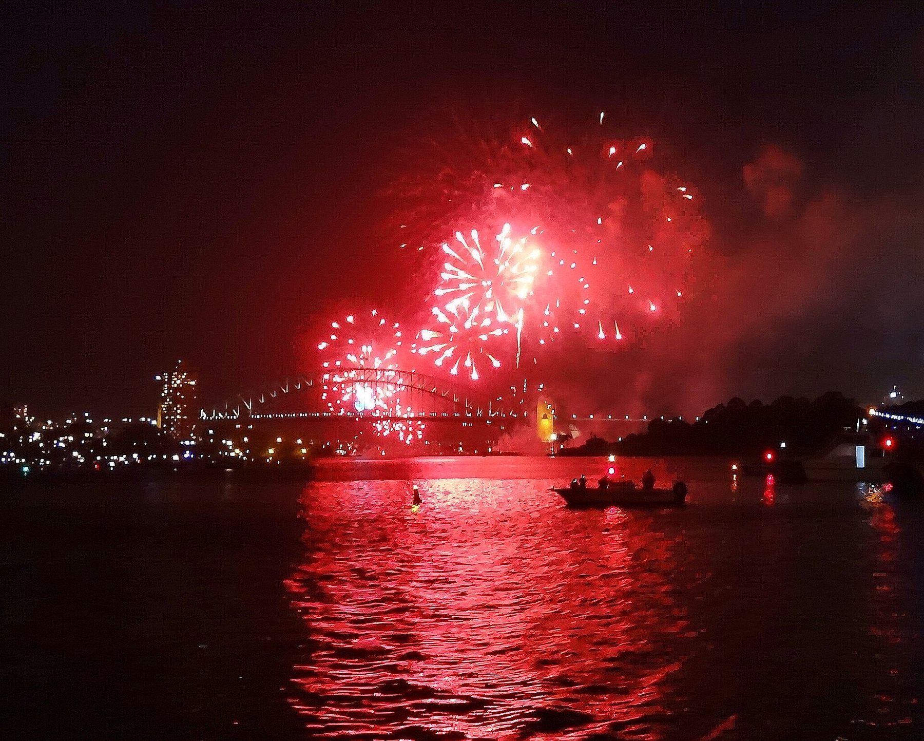 Red fireworks bursting over the Sydney Harbour Bridge at night, reflected in the water, with a small boat in silhouette and city lights along the shore.