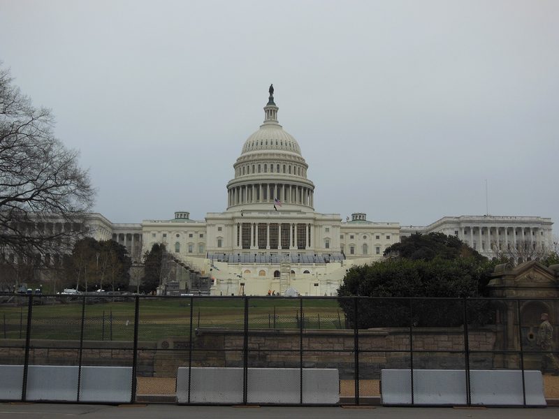 The United States Capitol under a grey sky, seen from behind a black security fence and a row of concrete barriers.