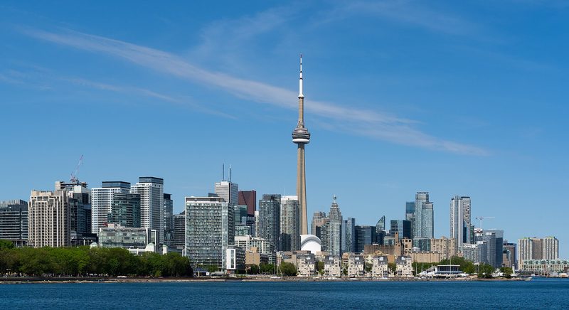 The Toronto skyline under a blue sky with thin streaks of cloud: the CN Tower rising above a long row of high-rise towers on the waterfront, seen across open blue water, with a line of green trees on the left shore