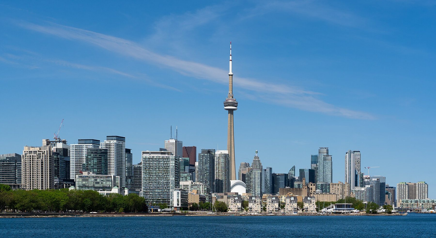 The Toronto skyline under a blue sky with thin streaks of cloud: the CN Tower rising above a long row of high-rise towers on the waterfront, seen across open blue water, with a line of green trees on the left shore