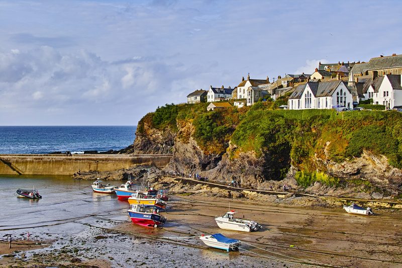 Small fishing boats resting on the sand of a harbour at low tide, below a green cliff topped with white and stone houses, with a breakwater and open sea to the left