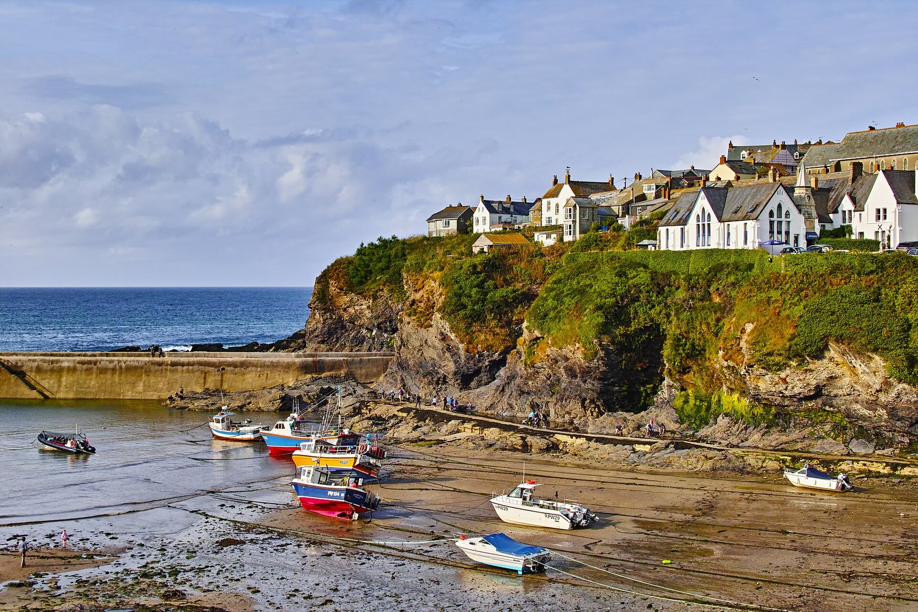 Small fishing boats resting on the sand of a harbour at low tide, below a green cliff topped with white and stone houses, with a breakwater and open sea to the left