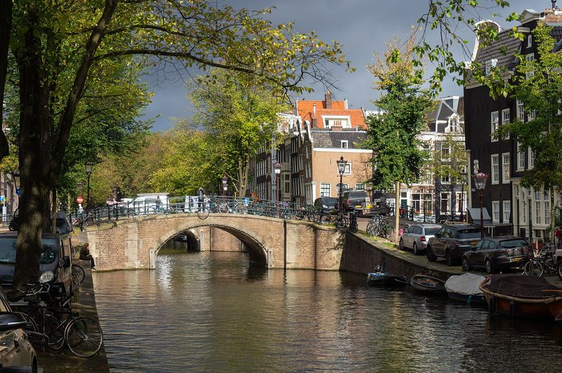 A brick arched bridge over a tree-lined canal, with bicycles along its railing, parked cars and small boats on the quay and tall canal houses behind under a dark sky