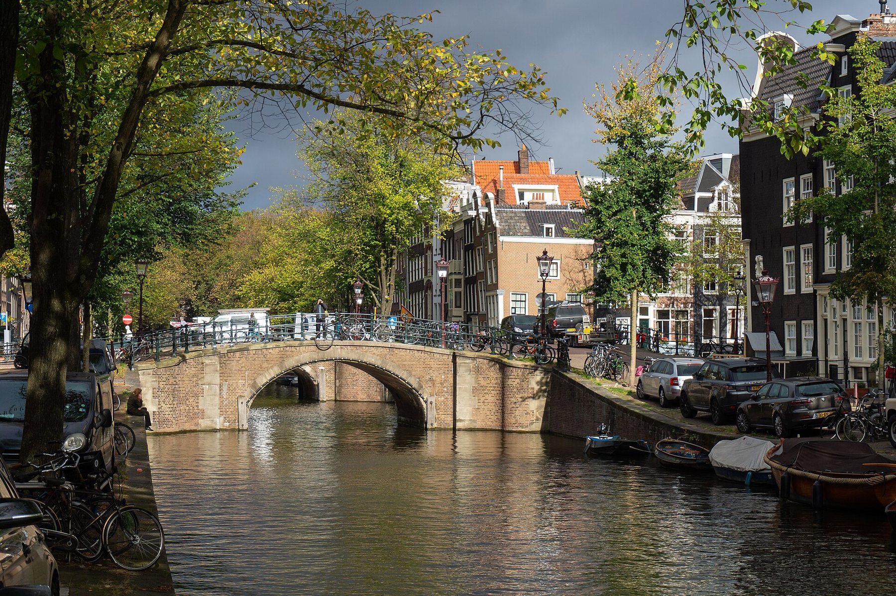 A brick arched bridge over a tree-lined canal, with bicycles along its railing, parked cars and small boats on the quay and tall canal houses behind under a dark sky