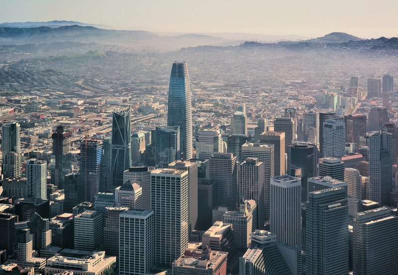 Downtown San Francisco from the air: office towers in haze, the tall rounded Salesforce Tower at the centre, hills behind