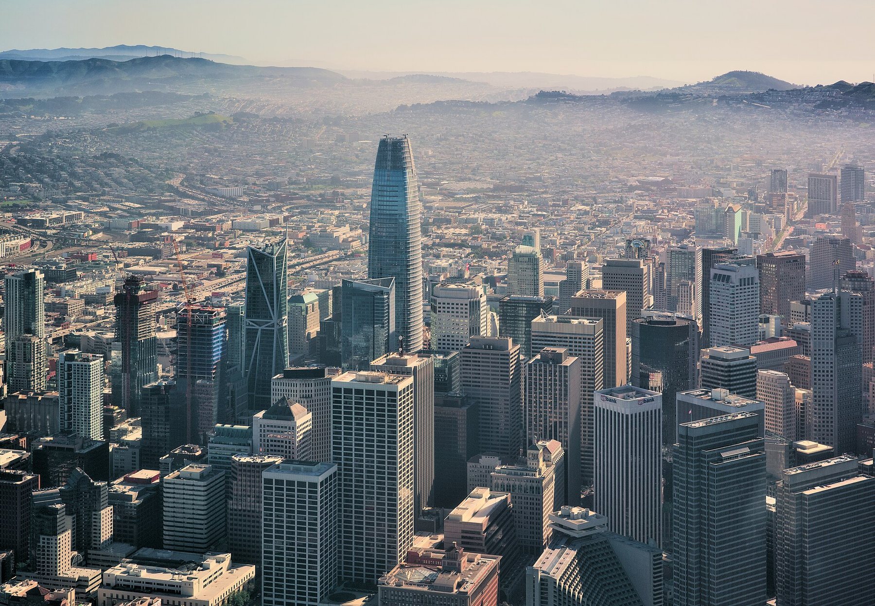 Downtown San Francisco from the air: office towers in haze, the tall rounded Salesforce Tower at the centre, hills behind