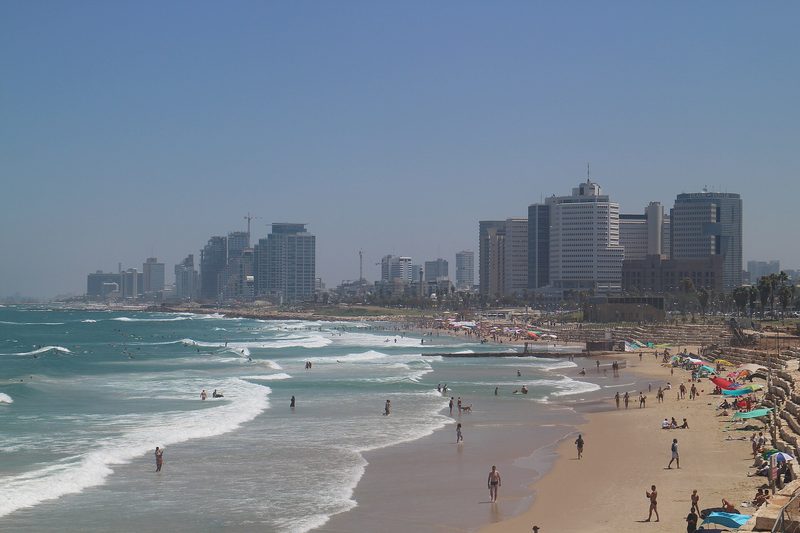 The Tel Aviv seafront on a clear day: waves breaking on a sandy beach with bathers and sun umbrellas, and high-rise towers lining the shore under a blue sky.