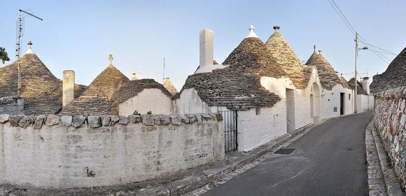A quiet lane in Alberobello lined with trulli: small whitewashed stone houses with cone-shaped roofs of grey stacked stone, behind a low dry-stone wall under a clear sky
