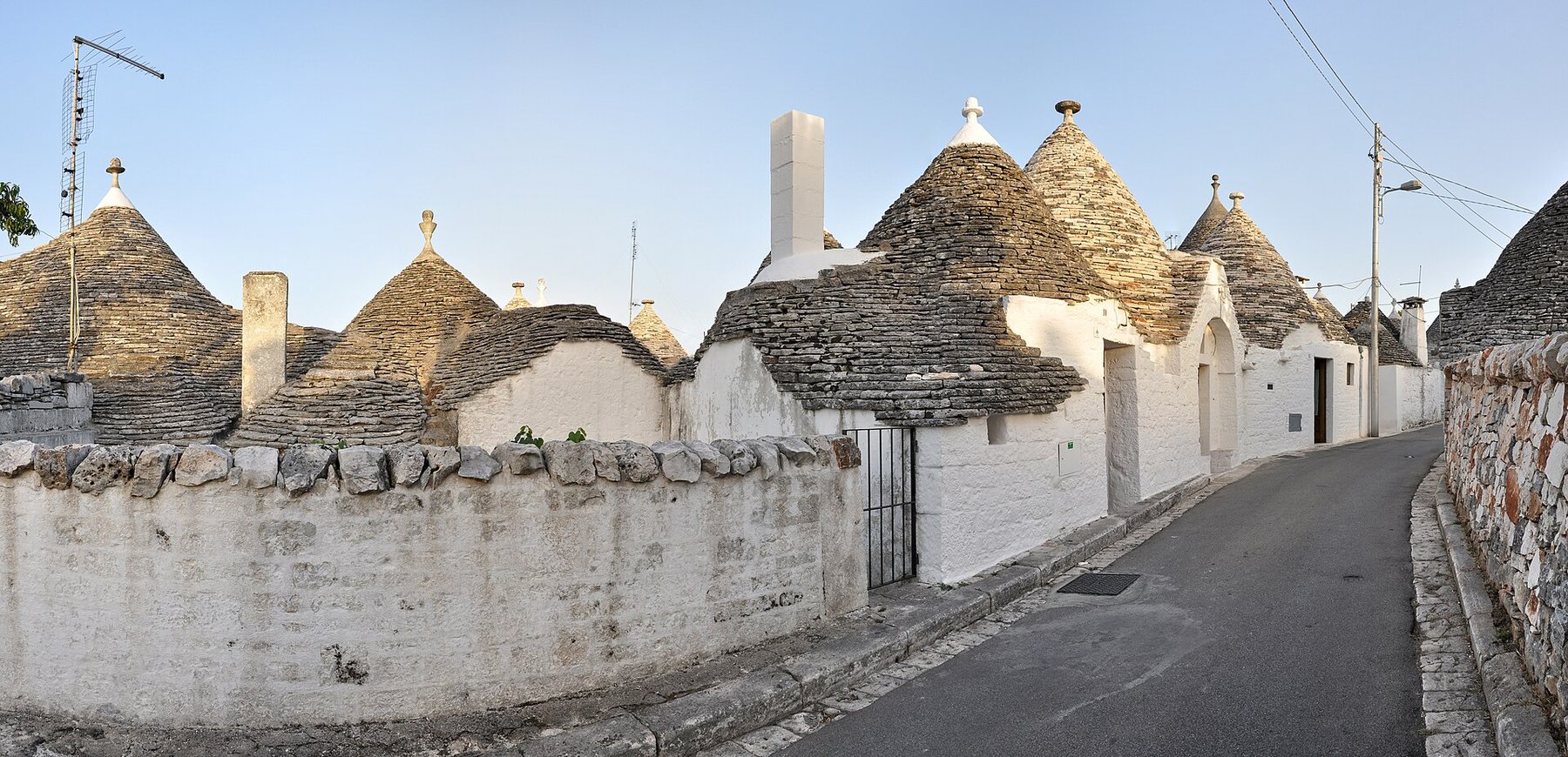 A quiet lane in Alberobello lined with trulli: small whitewashed stone houses with cone-shaped roofs of grey stacked stone, behind a low dry-stone wall under a clear sky