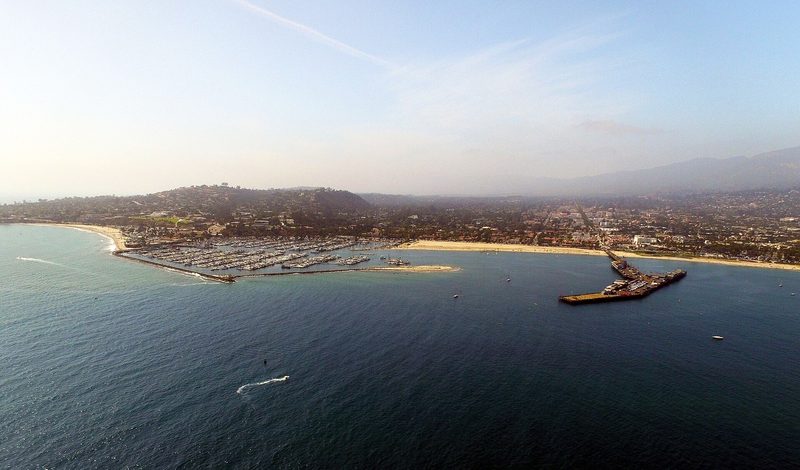 Aerial view from over the ocean of the Santa Barbara waterfront: a marina full of boats behind a breakwater, a long pier, a sandy beach and the city rising to hazy hills