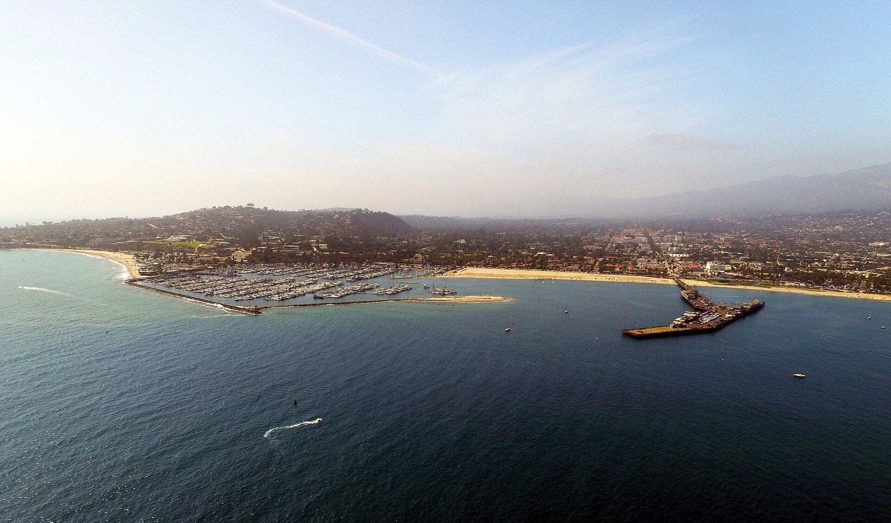 Aerial view from over the ocean of the Santa Barbara waterfront: a marina full of boats behind a breakwater, a long pier, a sandy beach and the city rising to hazy hills