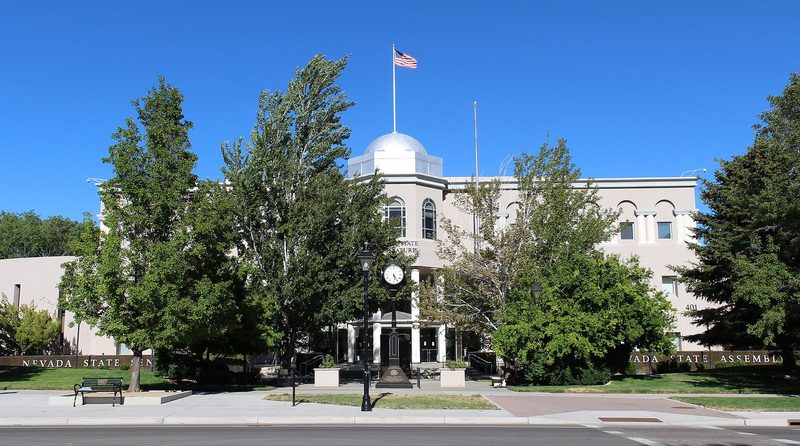 A pale two-story building with a small silver dome and an American flag on a pole, seen across a street through tall green trees under a clear blue sky, with a black street clock in front and low walls lettered Nevada State Senate and Nevada State Assembly